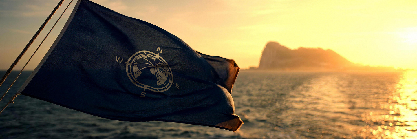 A navy house flag flying from a vessel’s stern at sunset, the Rock of Gibraltar beyond